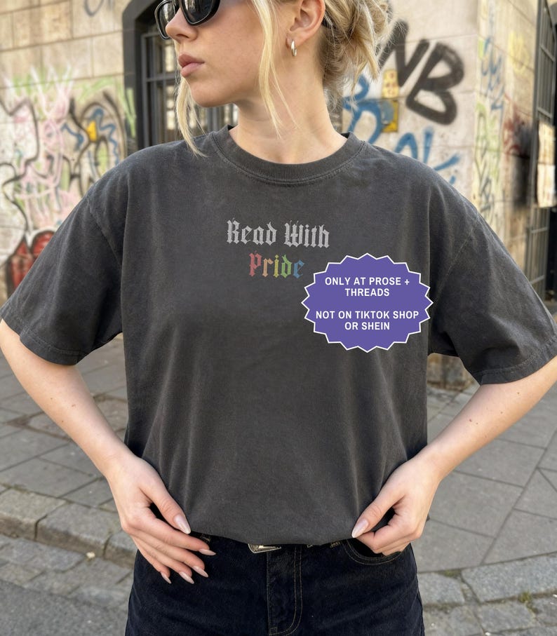 Pu&ograve; includere: Una t-shirt a maniche corte grigio antracite con il testo Read With Pride in un carattere gotico, con la parola Pride visualizzata nei colori dell'arcobaleno. Un grafico viola a forma di stella include il testo ONLY AT PROSE + THREADS NOT ON TIKTOK SHOP OR SHEIN.