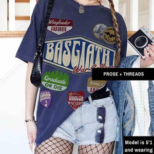 May include: A navy blue t-shirt with a vintage-style graphic design featuring the text "Basgaath War College" and various patches, including "Wingleader Favorite", "Graduate or Die", "Dragon Bonded", and "Three Survivor".