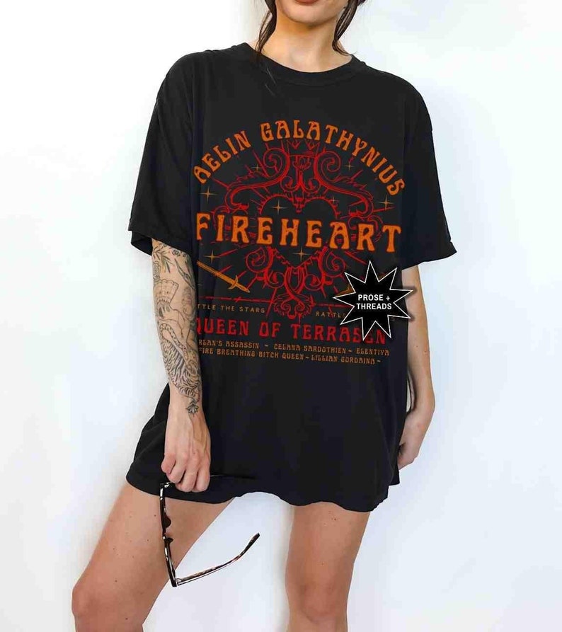 Pode incluir: T-shirt preto com o texto "AELIN GALATHYNIUS FIREHEART QUEEN OF TERRASEN" em laranja e vermelho. A camisa tem um design gr&aacute;fico com uma espada e estrelas. A camisa &eacute; usada por uma pessoa.