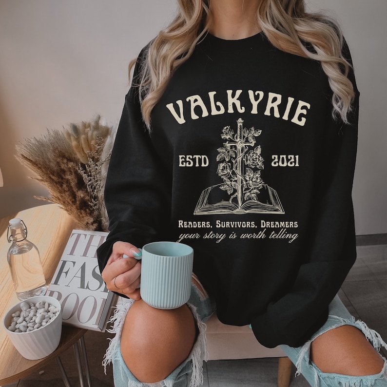 Może przedstawiać: Czarny sweter z białym nadrukiem graficznym przedstawiającym miecz przechodzący przez książkę z r&oacute;żami. Tekst brzmi "VALKYRIE ESTD 2021 READERS, SURVIVORS, DREAMERS your story is worth telling".