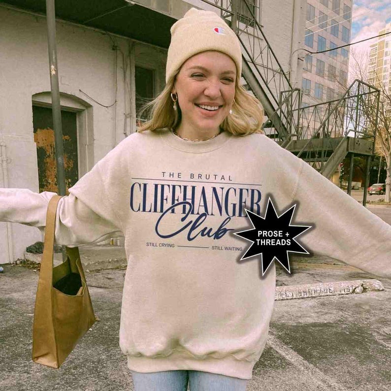 Puede incluir: Una sudadera beige con el texto "The Brutal Cliffhanger Club" en azul marino. El texto "Still Crying" y "Still Waiting" est&aacute; impreso en negro debajo del texto principal. Una estrella en blanco y negro con el texto "Prose + Threads" est&aacute; impresa en el lado derecho de la sudadera.