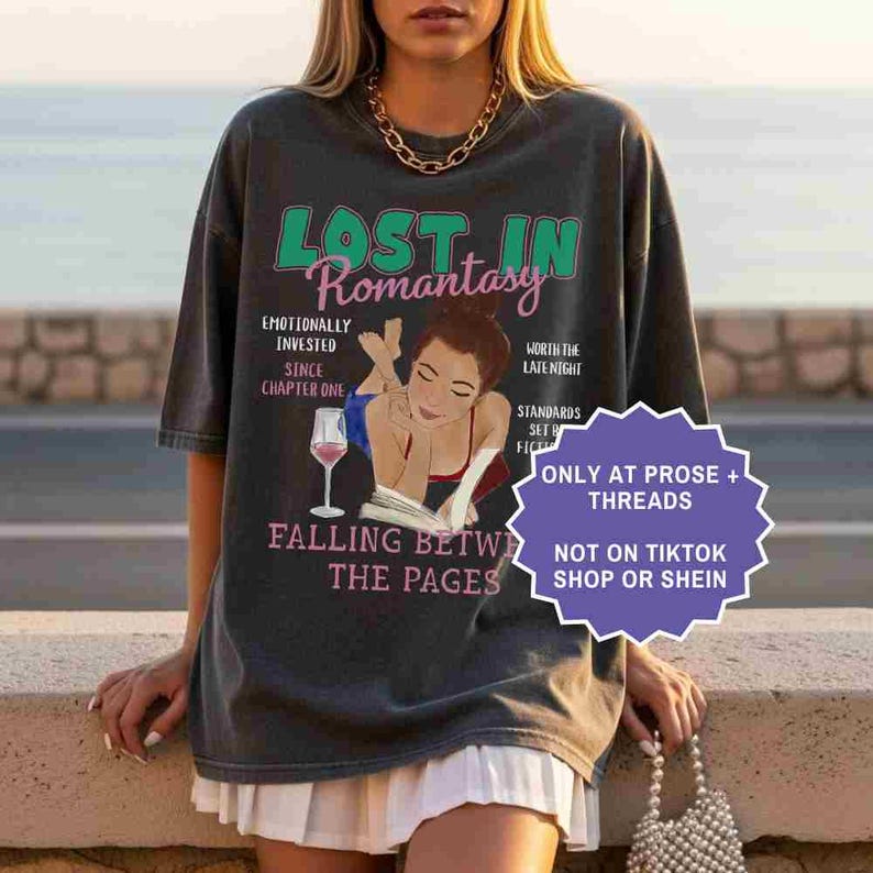 May include: Dark gray t-shirt with the text "LOST IN Romantasy" and a graphic of a woman reading a book with a glass of wine. Additional text includes "Emotionally Invested", "Since Chapter One", and "Falling Between the Pages".