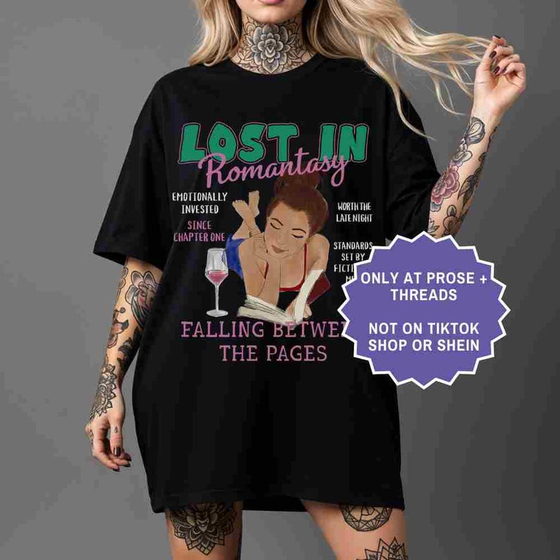 May include: Black t-shirt with the text "LOST IN Romantasy" in green and pink. The shirt features an illustration of a person reading a book with a glass of wine. Additional text includes "Emotionally Invested Since Chapter One".