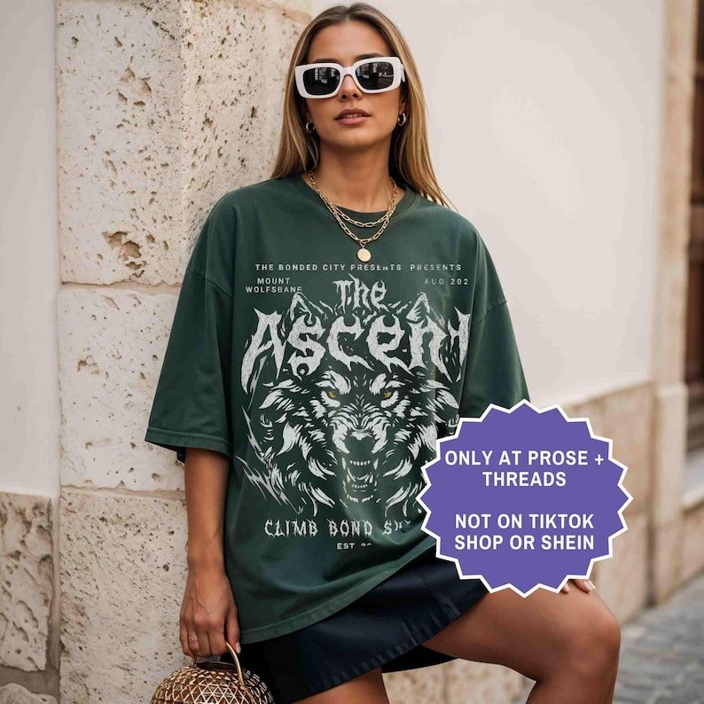 May include: Dark green oversized t-shirt with a wolf graphic and the text "The Ascent". The shirt also has the text "The Bonded City Presents" and "Climb Bond St". The shirt is worn with sunglasses and a gold necklace.