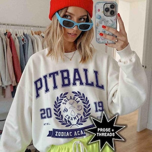 May include: White sweatshirt with a blue and white graphic print that says "Pitball" and "Solaris Champs 2019". The graphic also includes a wreath with a water drop, a flame, and a plant. The sweatshirt has a blue and white ribbon with the text "Zodiac Acai" on it.