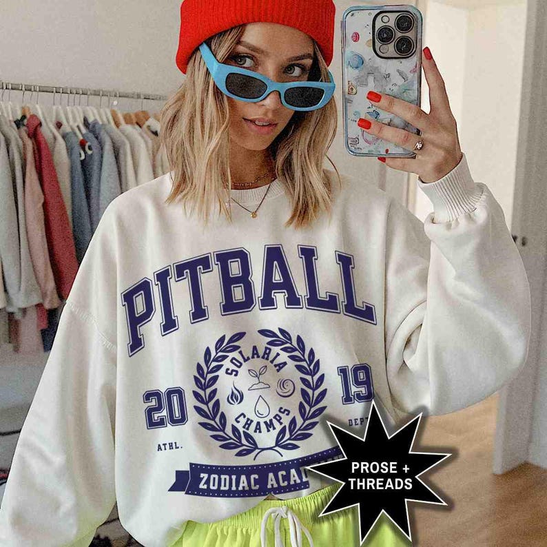 May include: White sweatshirt with a blue and white graphic print that says "Pitball" and "Solaris Champs 2019". The graphic also includes a wreath with a water drop, a flame, and a plant. The sweatshirt has a blue and white ribbon with the text "Zodiac Acai" on it.