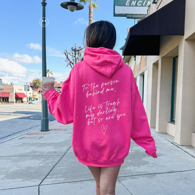 May include: A bright pink hoodie with white text on the back. The text reads, "To the person behind me, Life is tough my darling, but so are you" with a heart symbol. The hoodie is worn by a person walking on a sidewalk.