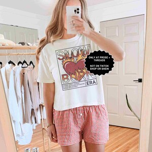May include: White t-shirt with a graphic of a heart on fire and the words "SLOW BURN ERA". The shirt is paired with red and white checkered shorts. The image also includes text that says "ONLY AT PROSE + THREADS" and "NOT ON TIKTOK SHOP OR SHEIN".