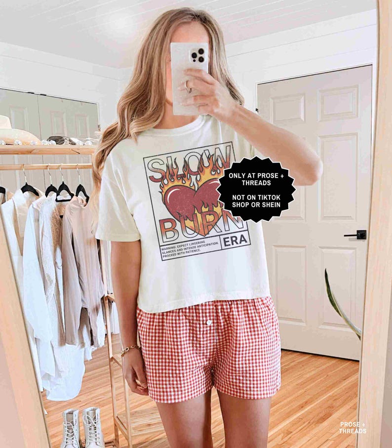 May include: White t-shirt with a graphic of a heart on fire and the words "SLOW BURN ERA". The shirt is paired with red and white checkered shorts. The image also includes text that says "ONLY AT PROSE + THREADS" and "NOT ON TIKTOK SHOP OR SHEIN".