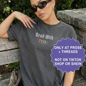 Pu&ograve; includere: Una t-shirt a maniche corte grigio antracite con la scritta Read With Pride in carattere gotico, con la parola Pride visualizzata nei colori dell'arcobaleno. La maglietta &egrave; indossata con occhiali da sole neri e una borsa a tracolla marrone.