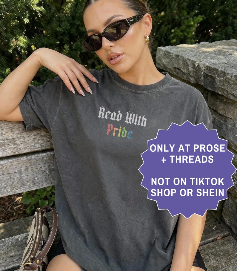 Pu&ograve; includere: Una t-shirt a maniche corte grigio antracite con la scritta Read With Pride in carattere gotico, con la parola Pride visualizzata nei colori dell'arcobaleno. La maglietta &egrave; indossata con occhiali da sole neri e una borsa a tracolla marrone.