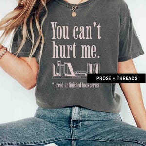 May include: A dark gray t-shirt with the text "You can't hurt me." in pink lettering. Below the text is a graphic of a bookshelf with books and the text "*I read unfinished book series" in pink lettering. The text "PROSE + THREADS" is in white lettering on a black background at the bottom right of the image. The t-shirt is labeled "SMALL".