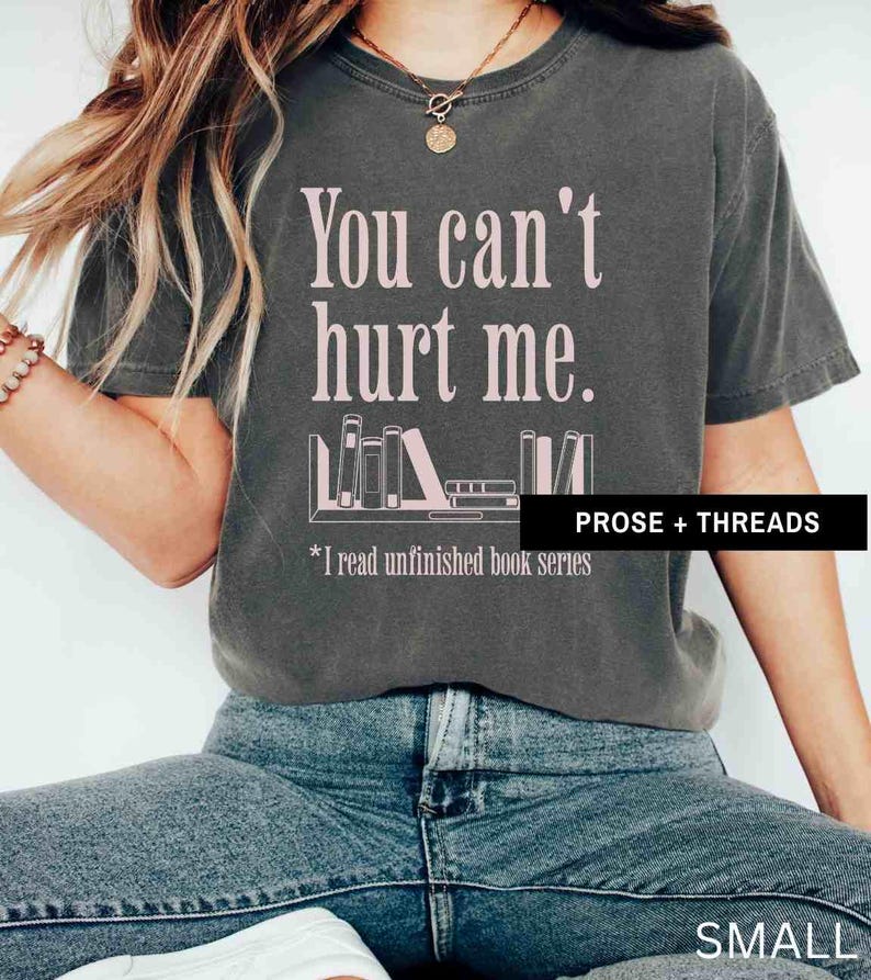 May include: A dark gray t-shirt with the text "You can't hurt me." in pink lettering. Below the text is a graphic of a bookshelf with books and the text "*I read unfinished book series" in pink lettering. The text "PROSE + THREADS" is in white lettering on a black background at the bottom right of the image. The t-shirt is labeled "SMALL".