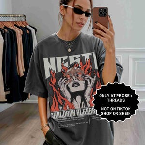 May include: A dark gray graphic t-shirt with the word "MERTI" in large white letters. The shirt features a graphic of a woman with flames and the text "CAULDRON BLESS6". The shirt is oversized and worn by a person.