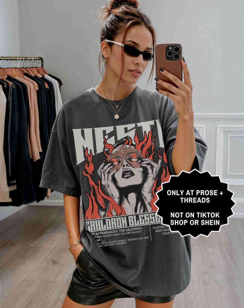 May include: A dark gray graphic t-shirt with the word "MERTI" in large white letters. The shirt features a graphic of a woman with flames and the text "CAULDRON BLESS6". The shirt is oversized and worn by a person.