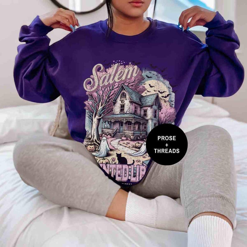 May include: Purple sweatshirt with a graphic print of a house with a spooky Halloween theme. The graphic includes the text "Salem Haunted" and features a black cat, ghosts, and pumpkins. The text "Prose + Threads" is also included in the graphic.