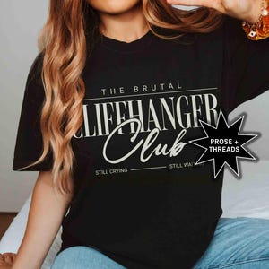 May include: Black t-shirt with white text that reads "The Brutal Cliffhanger Club" with the words "Still Crying" and "Still Waiting" underneath. A star with the words "Prose + Threads" is in the upper right corner.