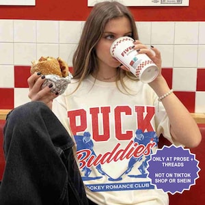 May include: A person wearing a cream-colored t-shirt with the words "PUCK Buddies" in red and blue, with hockey player graphics. The person is holding a burger in foil and a Five Guys cup. The shirt also says "Hockey Romance Club".