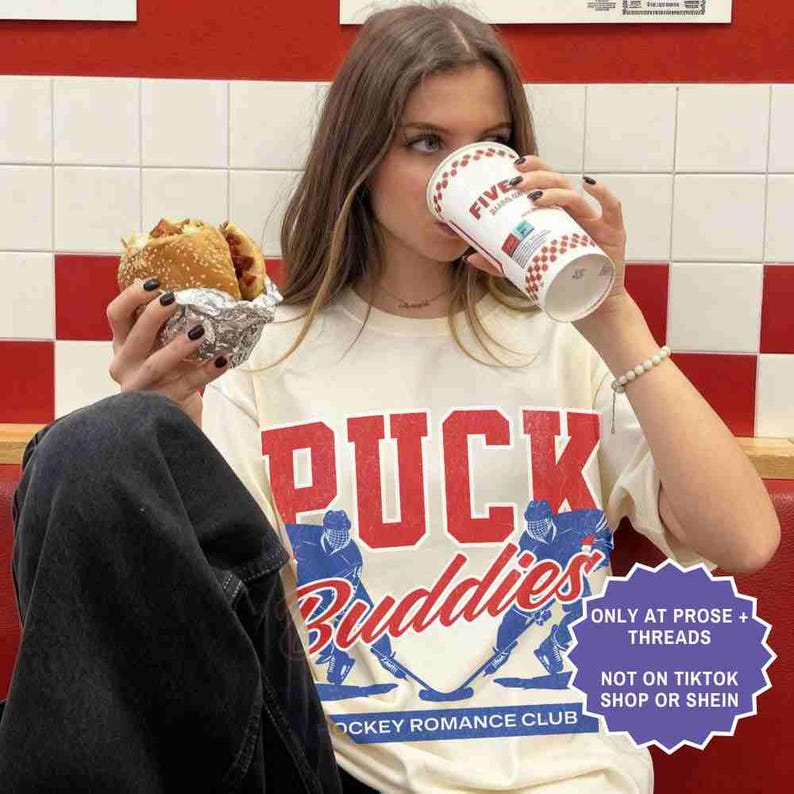 May include: A person wearing a cream-colored t-shirt with the words "PUCK Buddies" in red and blue, with hockey player graphics. The person is holding a burger in foil and a Five Guys cup. The shirt also says "Hockey Romance Club".