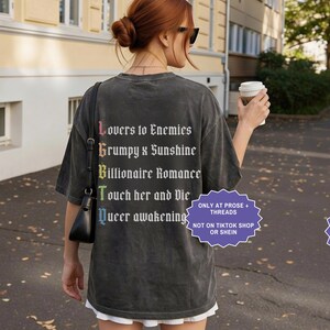 Pu&ograve; includere: Una t-shirt a maniche corte grigio antracite con testo bianco sul retro che recita Lovers to Enemies, Grumpy x Sunshine, Billionaire Romance, Touch her and Die e Queer awakening, con le lettere iniziali in un gradiente arcobaleno.