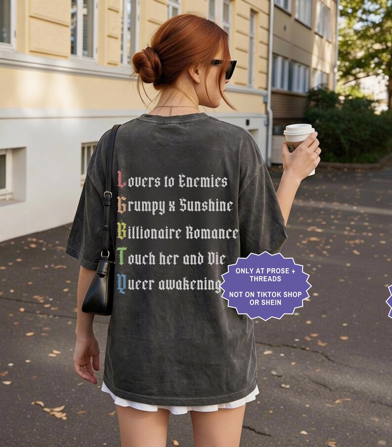 Pu&ograve; includere: Una t-shirt a maniche corte grigio antracite con testo bianco sul retro che recita Lovers to Enemies, Grumpy x Sunshine, Billionaire Romance, Touch her and Die e Queer awakening, con le lettere iniziali in un gradiente arcobaleno.