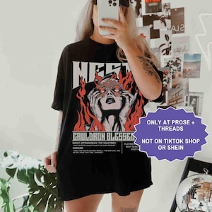 May include: Black t-shirt with a graphic of a woman's face surrounded by flames and the text "CAULDRON BLESSED". The shirt also has the text "ONLY AT PROSE + THREADS" and "NOT ON TIKTOK SHOP OR SHEIN".