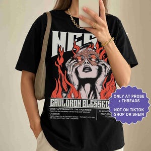 May include: Black t-shirt with a graphic of a woman with a mask and flames, the word "NER" above the graphic, and text that reads "CAULDRON BLESSED". The shirt also has the text "ONLY AT PROSE + THREADS" and "NOT ON TIKTOK SHOP OR SHEIN".