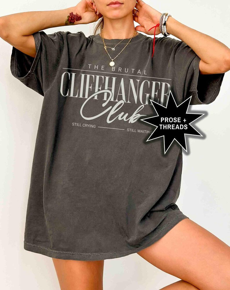 May include: A dark gray oversized t-shirt with the text "The Brutal Cliffhanger Club" printed on the front. The text is in white and has a vintage look. The shirt also has the text "Still Crying" and "Still Waiting" printed below the main text. A black star with the text "Prose + Threads" is printed on the right side of the shirt.