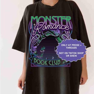 Op de afbeelding: Zwart T-shirt met een grafisch ontwerp met de tekst "MONSTER Romance BOOK CLUB 2". Het ontwerp bevat een silhouet van een wezen en decoratieve krullen in groen, paars en blauw. Het shirt heeft ook de tekst "ONLY AT PROSE + THREADS" en "NOT ON TIKTOK SHOP OR SHEIN".