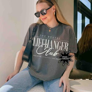 May include: A gray t-shirt with the text "The Brutal Lifehanger Club" printed on the front. The text is in white and has a distressed look. The shirt is being worn by a person with long brown hair and black sunglasses. The person is sitting on a white chair. The shirt also has the text "Still Crying" and "Still Waiting" printed on the bottom.