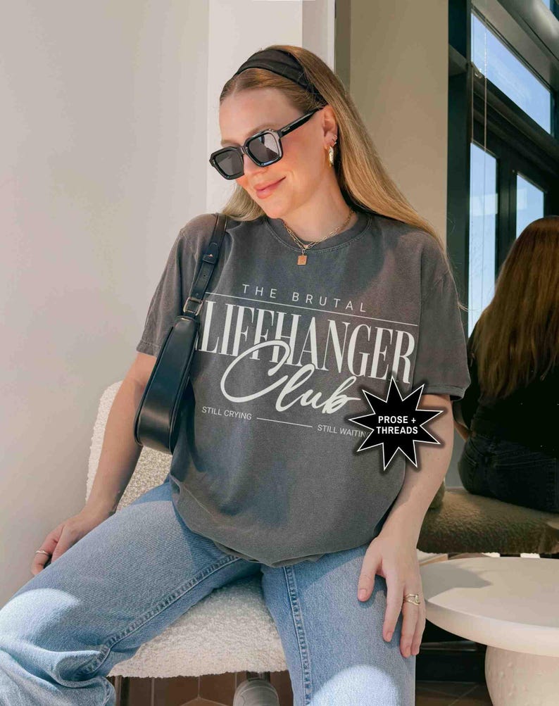May include: A gray t-shirt with the text "The Brutal Lifehanger Club" printed on the front. The text is in white and has a distressed look. The shirt is being worn by a person with long brown hair and black sunglasses. The person is sitting on a white chair. The shirt also has the text "Still Crying" and "Still Waiting" printed on the bottom.