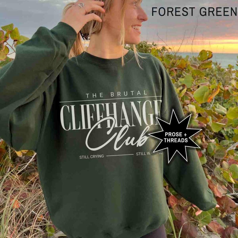 Puede incluir: Una sudadera de color verde bosque con el texto "The Brutal Cliffhanger Club" en blanco. El texto "Still Crying" y "Still Waiting" se encuentra debajo del texto principal. Una estrella negra con el texto "Prose + Threads" se encuentra en la esquina superior derecha.