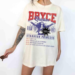 May include: An oversized cream-coloured t-shirt with a vintage-style graphic print featuring a woman with long dark hair, the text "Bryce Quinlan", and the words "Starborn Princess". The graphic also includes the text "Prose + Threads" and a list of dates and phrases.