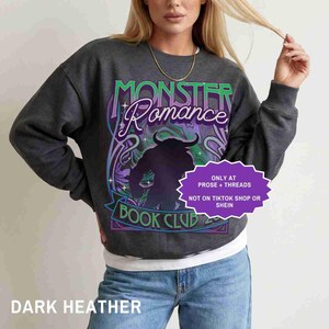 May include: Dark heather gray sweatshirt with the text "Monster Romance Book Club" in green and purple. The design includes a silhouette of a monster and decorative floral elements. The sweatshirt is a casual style.