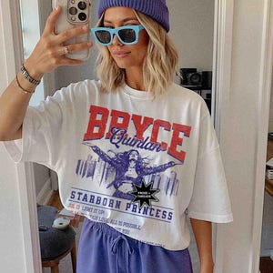 May include: White t-shirt featuring a graphic print of a woman with the text "Bryce Quinlan" and "Starborn Princess". The graphic is in red, blue, and black. The shirt has a crew neck and short sleeves.