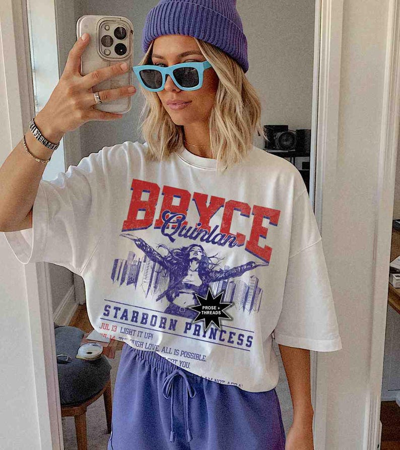 May include: White t-shirt featuring a graphic print of a woman with the text "Bryce Quinlan" and "Starborn Princess". The graphic is in red, blue, and black. The shirt has a crew neck and short sleeves.