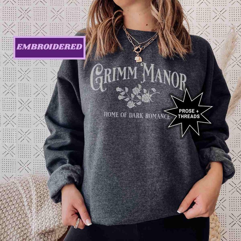 May include: Dark grey embroidered sweatshirt featuring the text "Grimm Manor, Home of Dark Romance". The design includes floral elements. The sweatshirt is a long-sleeved crew neck.
