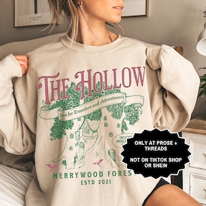 May include: Beige sweatshirt featuring a pink and green graphic with the words "The Hollow". The design includes a tree with a house, the text "Inn for Travelers and Adventurers", and "Merrywood Fores ESTD 2021".