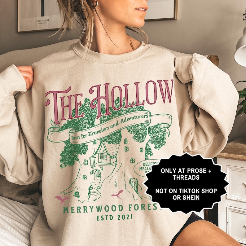 May include: Beige sweatshirt featuring a pink and green graphic with the words "The Hollow". The design includes a tree with a house, the text "Inn for Travelers and Adventurers", and "Merrywood Fores ESTD 2021".