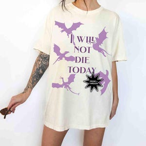 May include: A white oversized t-shirt with a purple graphic of dragons and the text "I WILL NOT DIE TODAY". The shirt has a black star-shaped pin with the text "PROSE + THREADS".