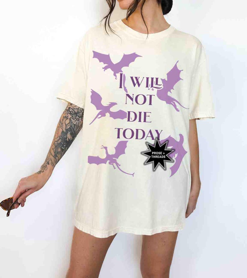 May include: A white oversized t-shirt with a purple graphic of dragons and the text "I WILL NOT DIE TODAY". The shirt has a black star-shaped pin with the text "PROSE + THREADS".