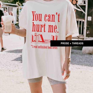 May include: A white t-shirt with red text that says "You can't hurt me."  The text is followed by a graphic of a bookshelf with books on it.  The text below the graphic says "*I read unfinished book series".  The text "PROSE + THREADS" is in the bottom right corner of the image.