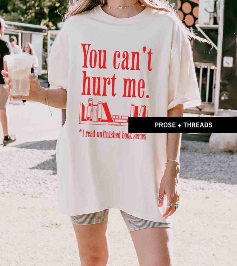 May include: A white t-shirt with red text that says "You can't hurt me."  The text is followed by a graphic of a bookshelf with books on it.  The text below the graphic says "*I read unfinished book series".  The text "PROSE + THREADS" is in the bottom right corner of the image.