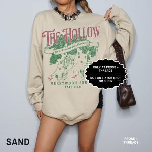 May include: A sand-coloured sweatshirt featuring a burgundy graphic that reads "The Hollow". The design includes a tree with a house, the text "Inn for Travelers and Adventurers," and "Merrywood For Estd 2021." The sweatshirt has long sleeves.