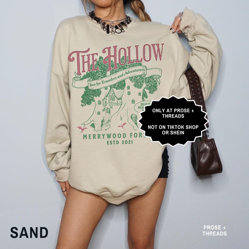 May include: A sand-coloured sweatshirt featuring a burgundy graphic that reads "The Hollow". The design includes a tree with a house, the text "Inn for Travelers and Adventurers," and "Merrywood For Estd 2021." The sweatshirt has long sleeves.