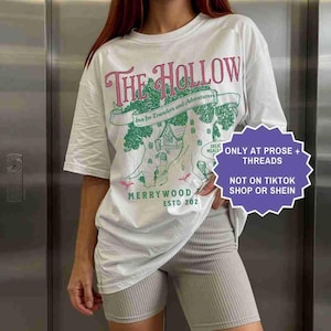 Pu&ograve; includere: T-shirt bianco con un design grafico con la scritta "THE HOLLOW" in rosa e un'illustrazione verde di un albero con una casa. &Egrave; presente anche il testo "Inn for Travelers and Adventurers". La maglietta ha anche la scritta "MERRYWOOD ESTD 202".