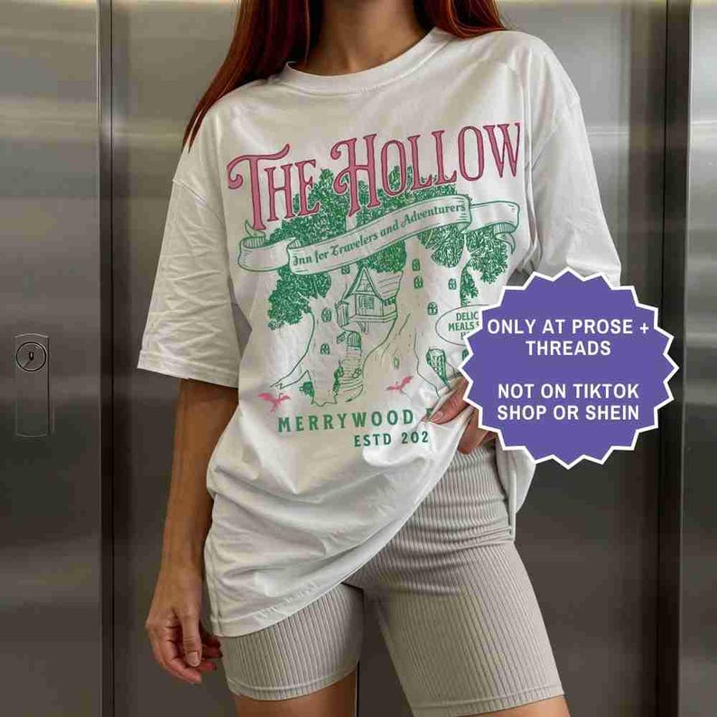 Pu&ograve; includere: T-shirt bianco con un design grafico con la scritta "THE HOLLOW" in rosa e un'illustrazione verde di un albero con una casa. &Egrave; presente anche il testo "Inn for Travelers and Adventurers". La maglietta ha anche la scritta "MERRYWOOD ESTD 202".