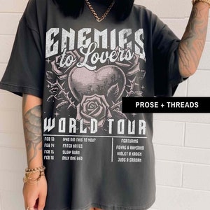 Pode incluir: Uma camiseta preta com uma estampa gr&aacute;fica de um cora&ccedil;&atilde;o com espinhos e rosas. O texto "Enemies to Lovers World Tour" est&aacute; impresso na camiseta. O texto "Featuring Feyre & Rhysand, Violet & Xaden, Jude & Cardan" est&aacute; impresso no lado direito da camiseta. O texto "Who did this to you? Fated Mates, Slow Burn, Only One Bed" est&aacute; impresso no lado esquerdo da camiseta.