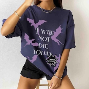 May include: A navy blue t-shirt with a graphic print of purple dragons and the text "I Will Not Die Today". The shirt has a relaxed fit and short sleeves. A small black starburst sticker with the text "Prose + Threads" is on the bottom right corner of the shirt.
