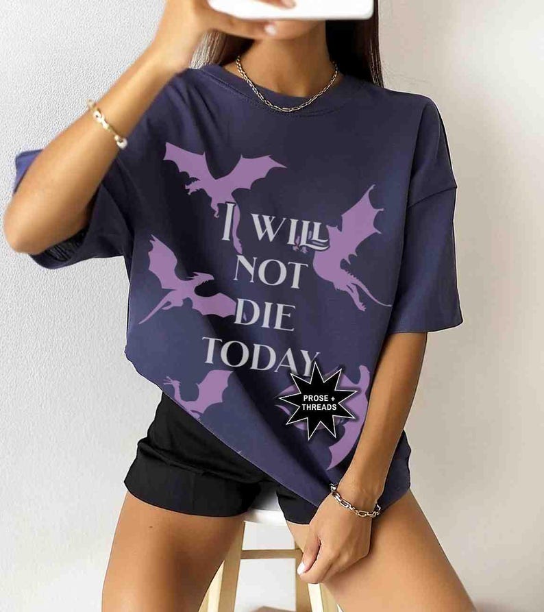 May include: A navy blue t-shirt with a graphic print of purple dragons and the text "I Will Not Die Today". The shirt has a relaxed fit and short sleeves. A small black starburst sticker with the text "Prose + Threads" is on the bottom right corner of the shirt.